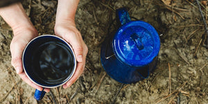 Woman holding a warm cup of coffee in camp with camping coffee pot nearby.